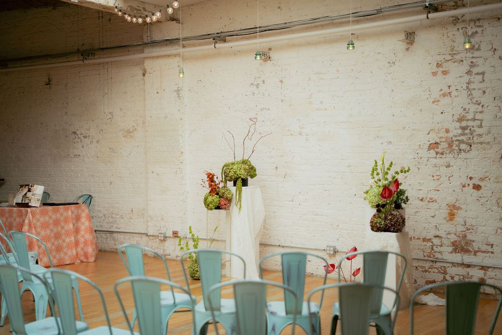 Rustic wedding venue interior with teal metal chairs and simple floral centerpieces against a distressed brick wall.