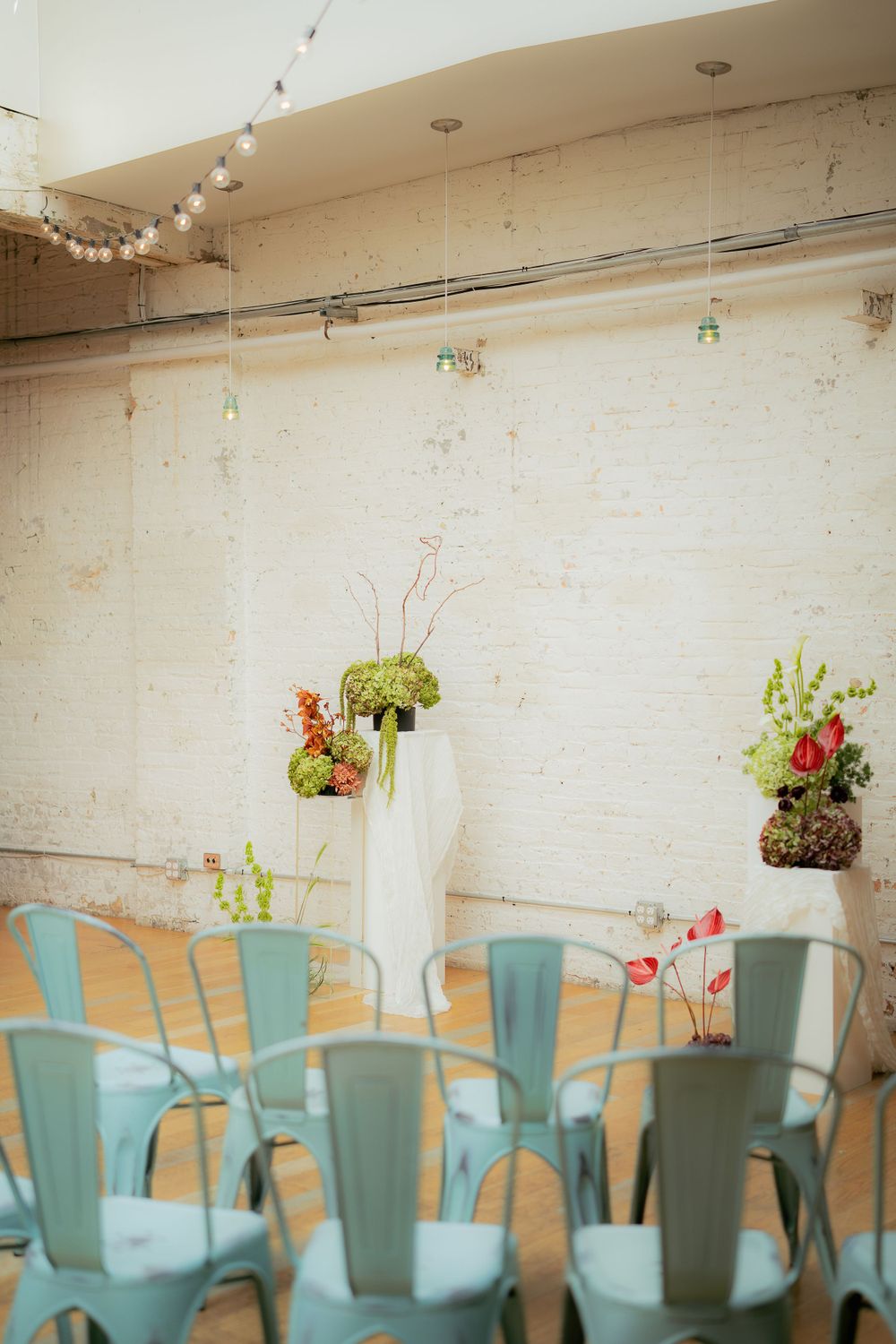 Light blue metal chairs arranged in rows with delicate floral arrangements in white vases against a rustic wall.