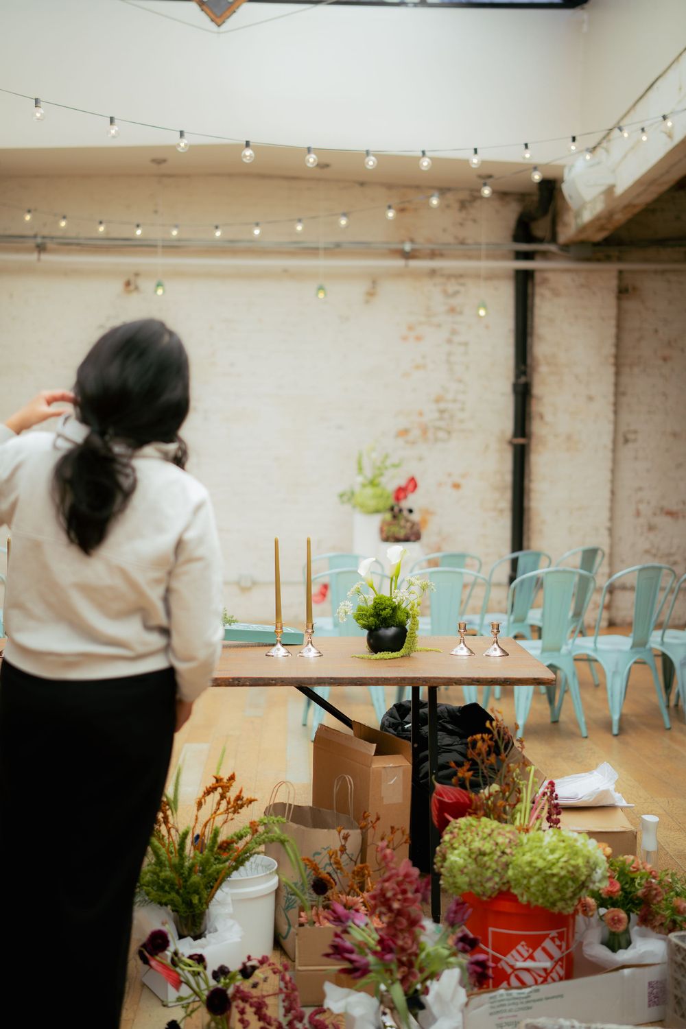 Indoor event space with string lights, turquoise chairs and floral arrangements being set up for a gathering.