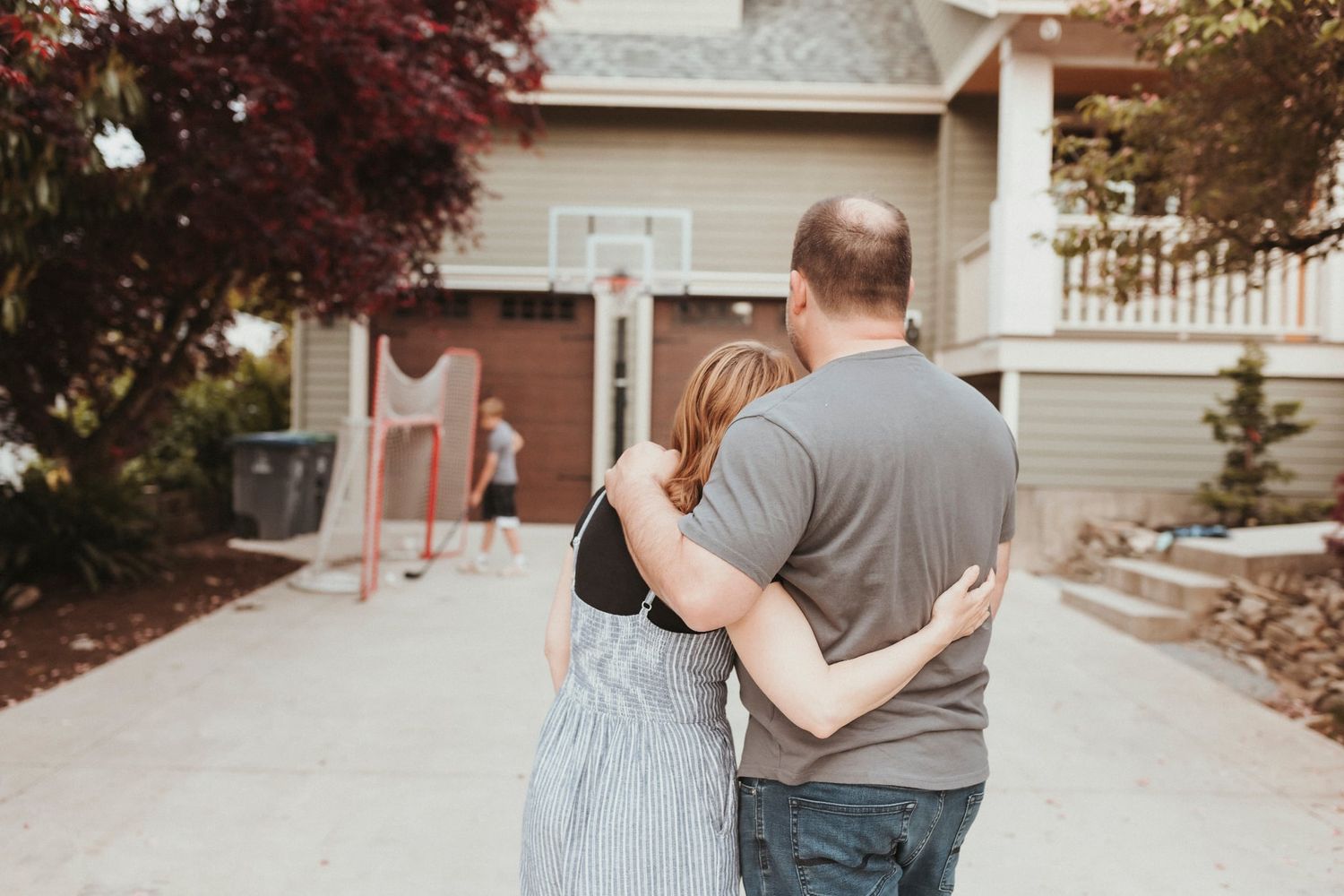 Husband and wife hug while their children play in the driveway of their home. Family photos in Surrey. Canada