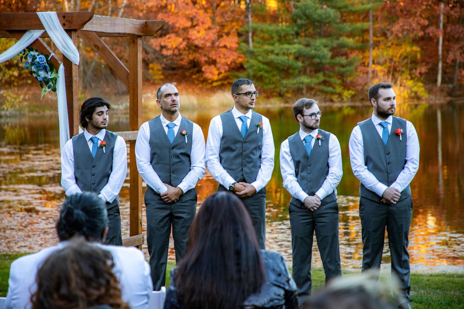 Groomsmen wearing matching gray vests stand in line at an outdoor fall wedding ceremony near a pond with autumn foliage.
