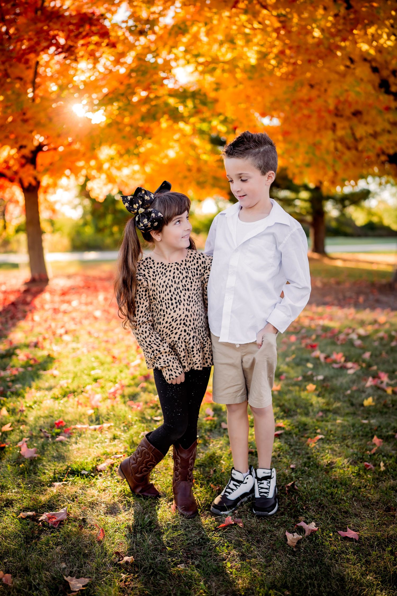 Two children pose together on grass beneath vibrant orange autumn trees during golden hour sunset lighting.