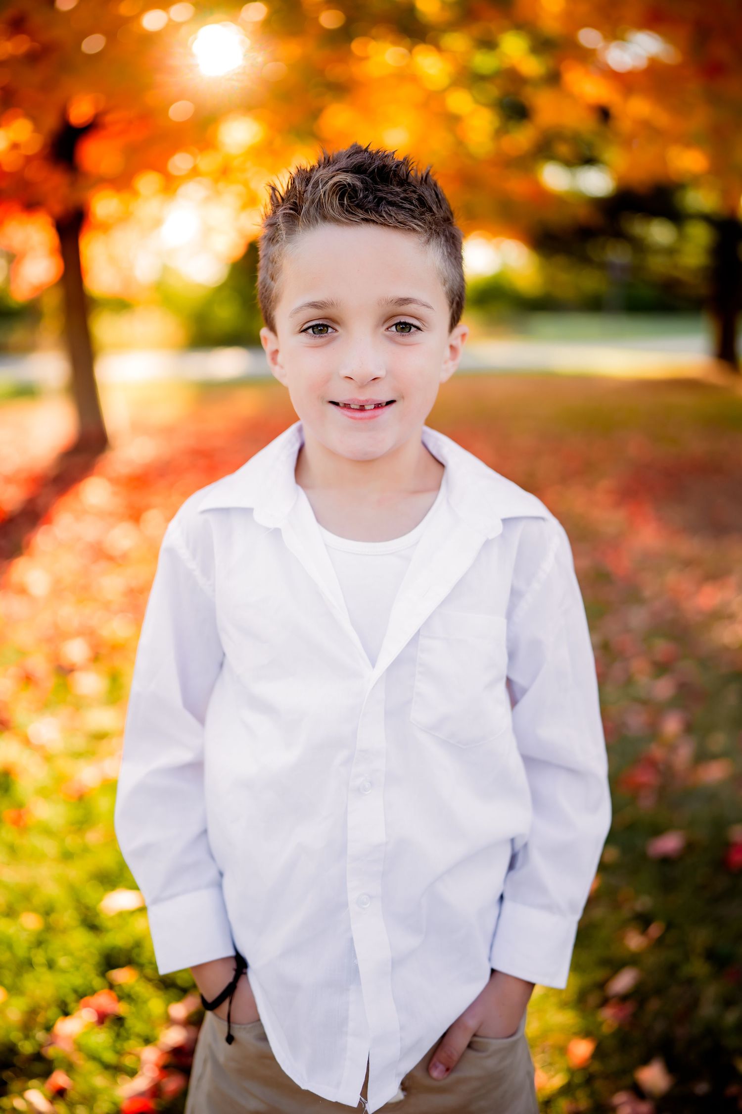 A child wearing white shirt and khaki pants stands outdoors with vibrant autumn foliage and golden sunlight in background.