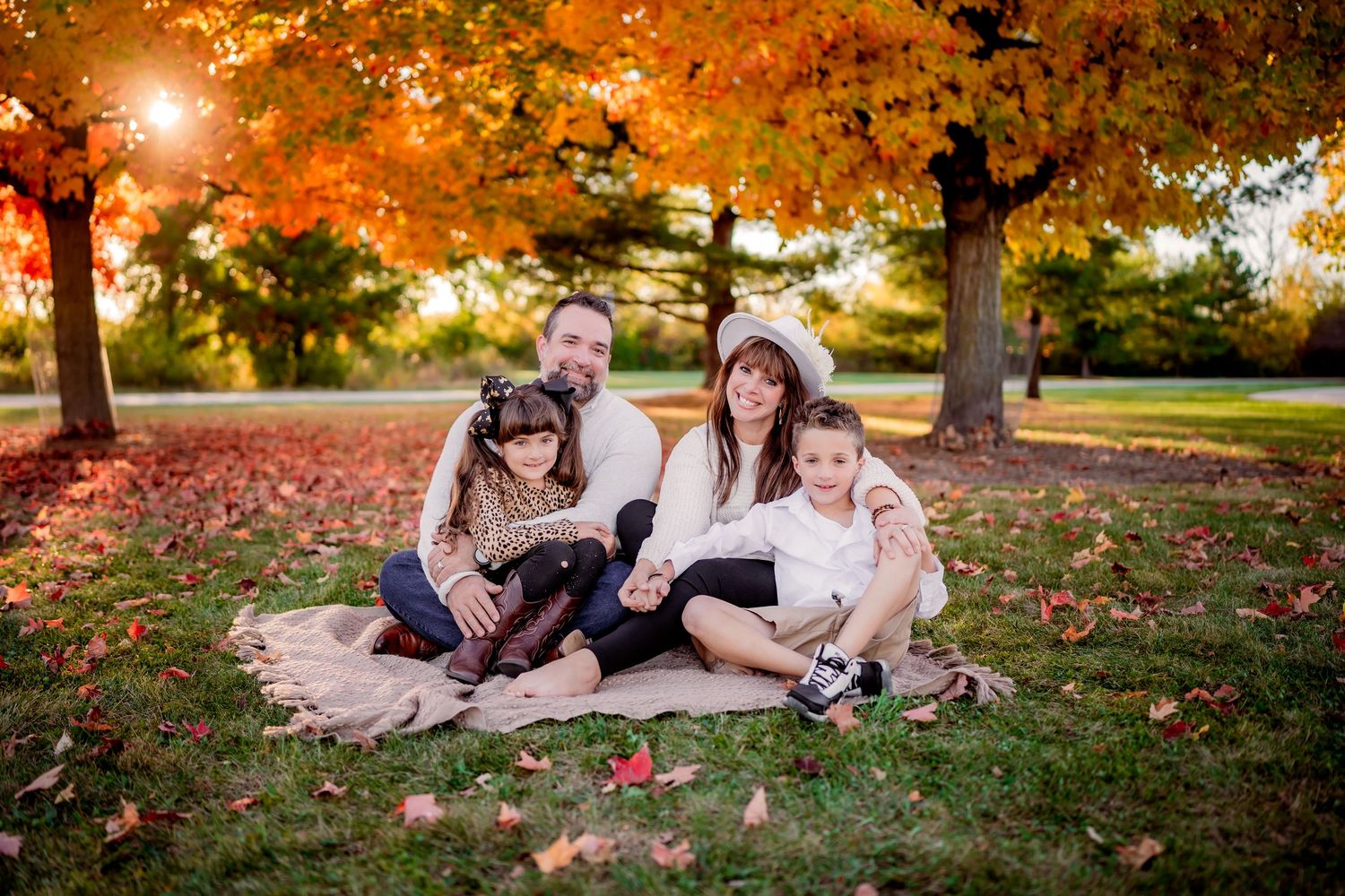 A family sits together on fall leaves during a warm autumn sunset in the park with golden light filtering through trees.
