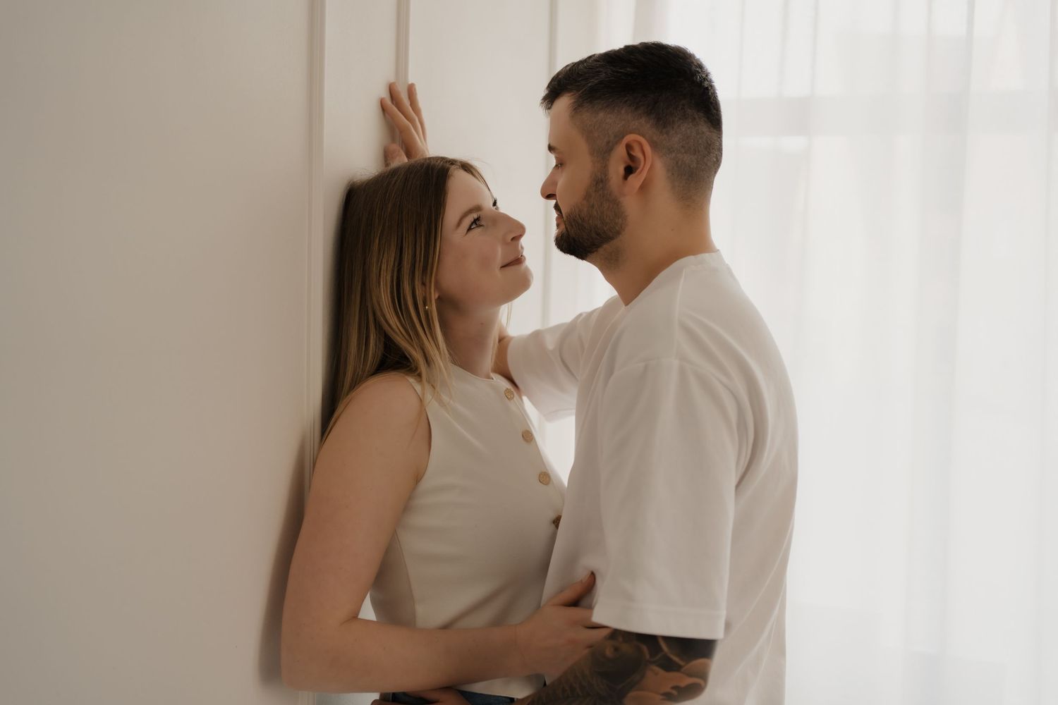 Two people in white outfits sharing an intimate moment against a white wall in soft natural light.
