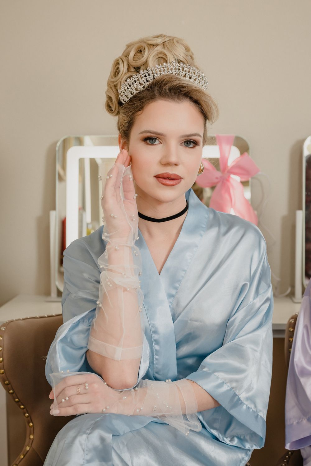 Elegant figure in blue satin robe wearing a crystal tiara and lace gloves poses in vintage-style boudoir setting.