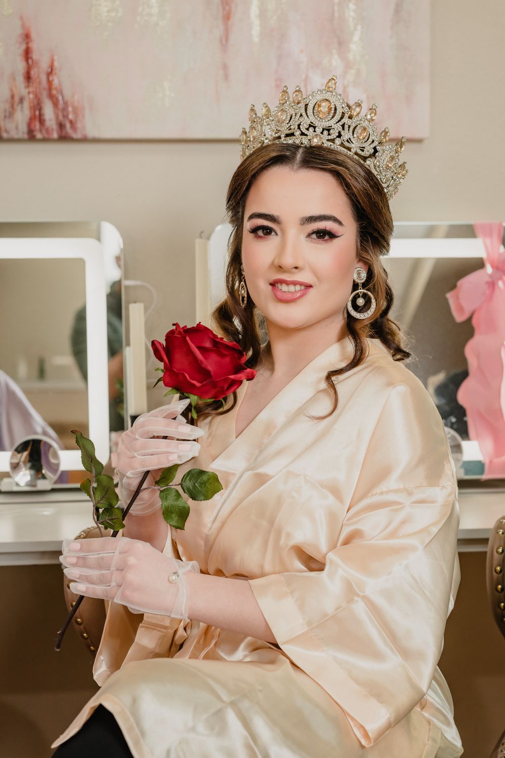 A golden-robed figure wearing an ornate crown holds a single red rose while sitting in front of vanity mirrors.