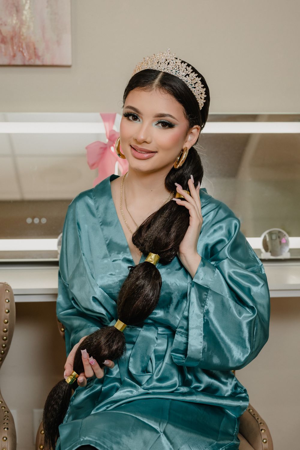 Person in teal satin robe wearing a crystal tiara poses with long braided hair.