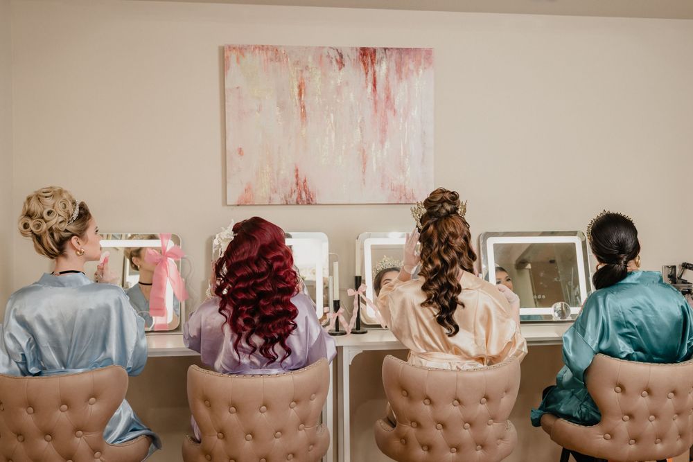 Four people in satin robes sit at a makeup counter with mirrors and a pink abstract painting on the wall.