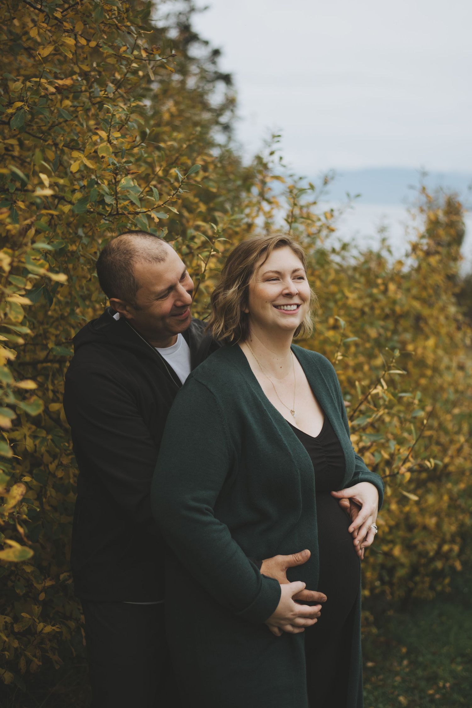 An expectant couple embraces outdoors in autumn with yellow leaves and foliage in the background.