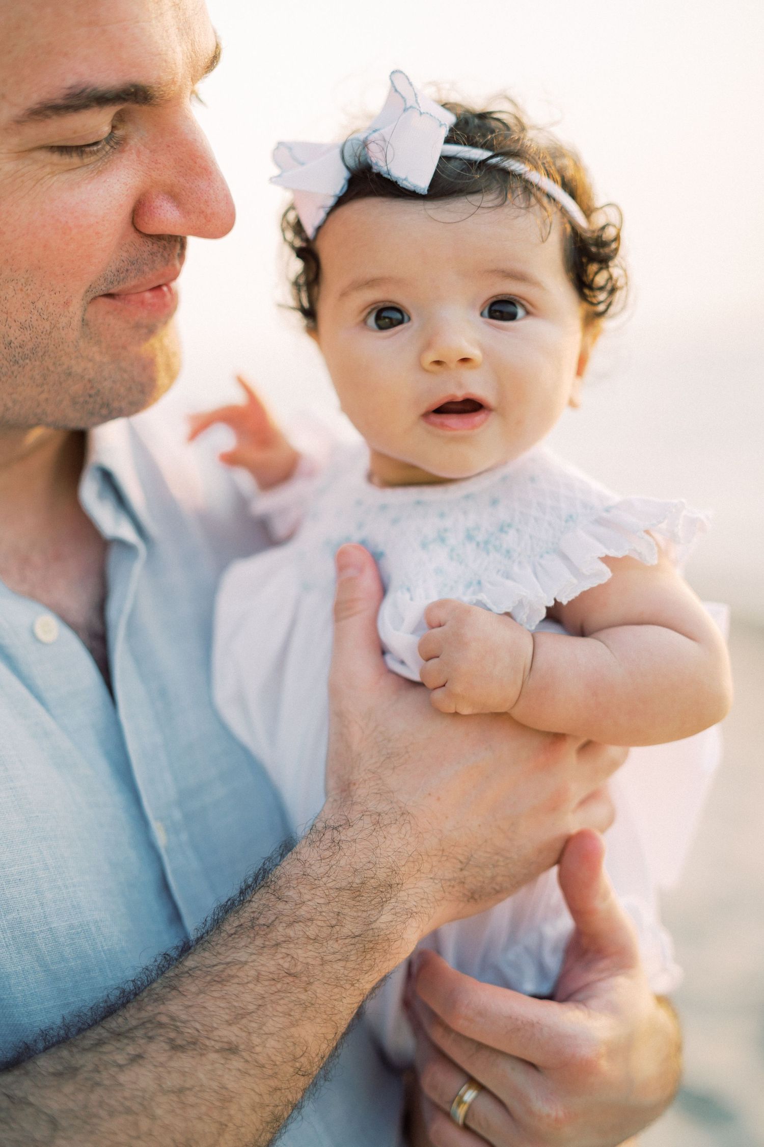 Tender moment between parent and baby in white lace dress with bow against soft light background.