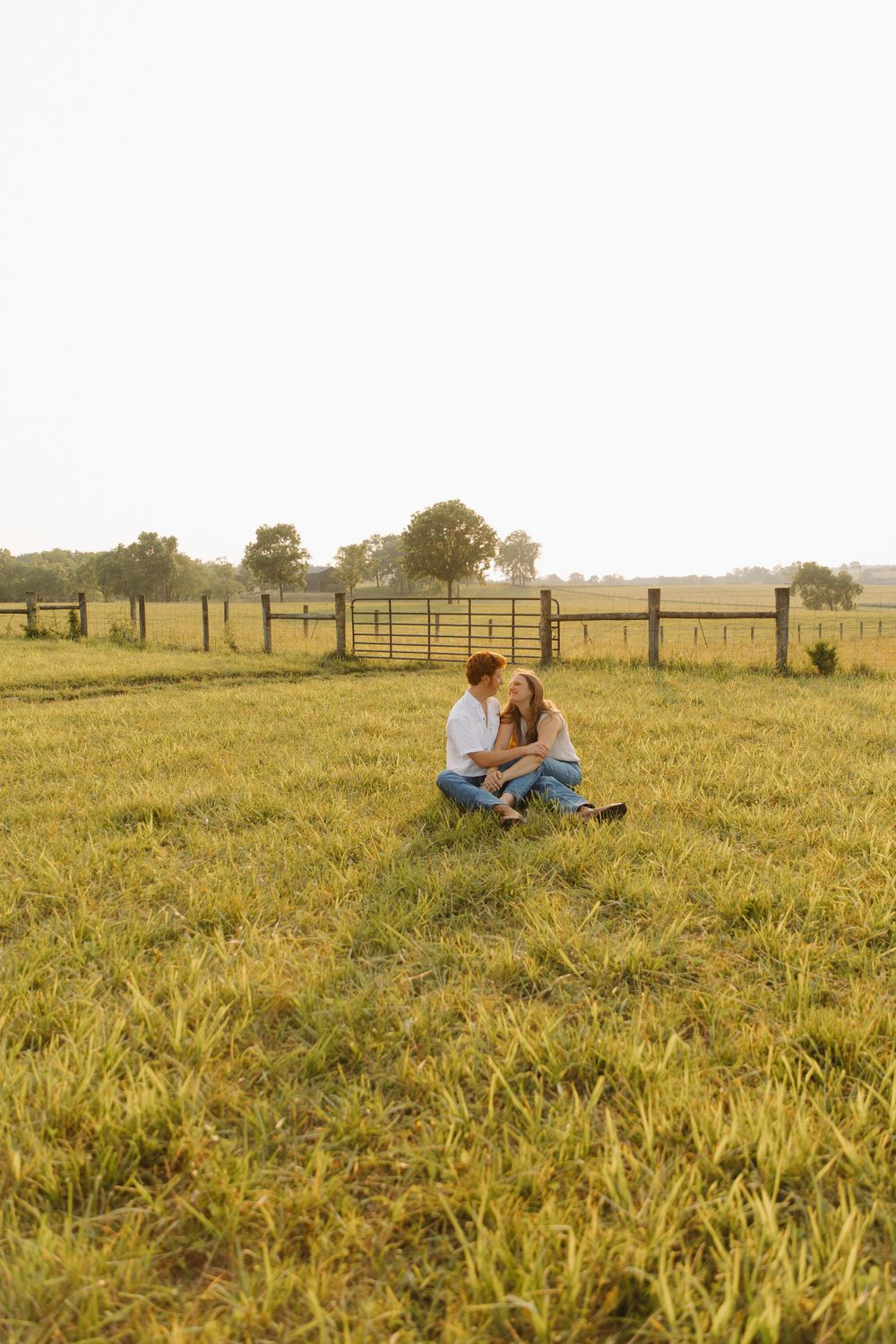 Couple sitting together in a sunlit meadow with wooden fences in the background in Lexington, Kentucky.