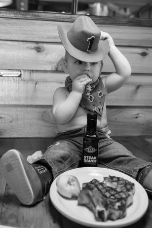 A black and white photo of a small child in western attire with a steak dinner.