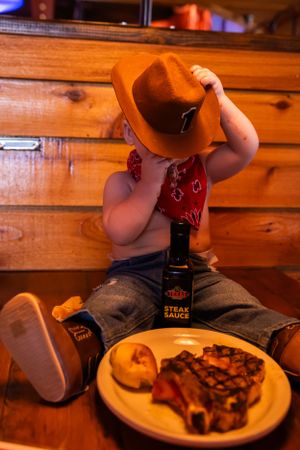A small child in cowboy attire plays with hat while seated at restaurant table.