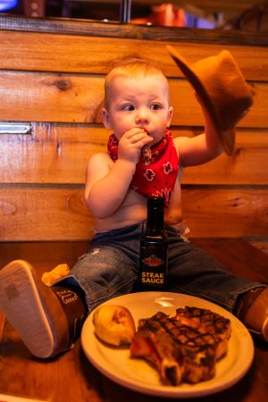 A toddler in western wear adjusts cowboy hat while seated with dinner plate.
