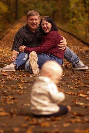 Couple sits on leaf-covered path while baby explores in foreground.