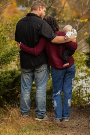 A couple in sweaters embraces while holding an infant on a fall nature trail.