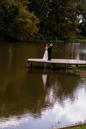 Wedding couple standing on wooden dock over serene lake surrounded by autumn trees.