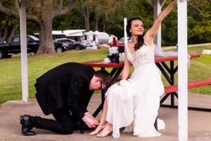 Candid moment of groom assisting bride with footwear during outdoor wedding photoshoot.