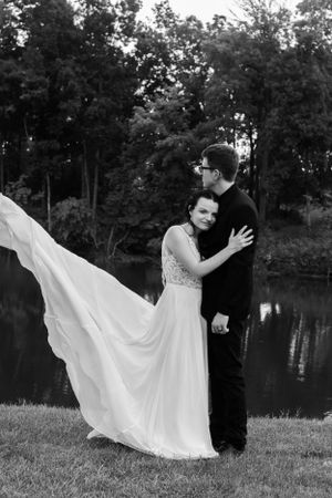 Black and white romantic portrait of newlyweds with bride's dress flowing in the wind by lake.