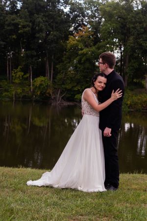 A romantic wedding couple embraces by a tranquil pond with trees in the background.