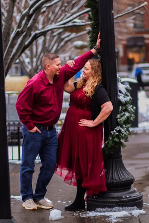 A couple in matching burgundy outfits poses playfully by a decorated lamp post in winter.
