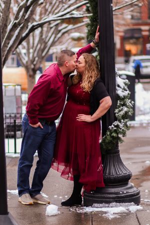 A couple in burgundy attire shares a romantic kiss by a lamp post during winter.