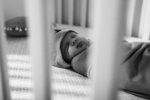 A black and white photograph of a sleeping baby viewed through crib bars.