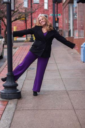Person in purple pants and black blazer striking playful pose on urban sidewalk.