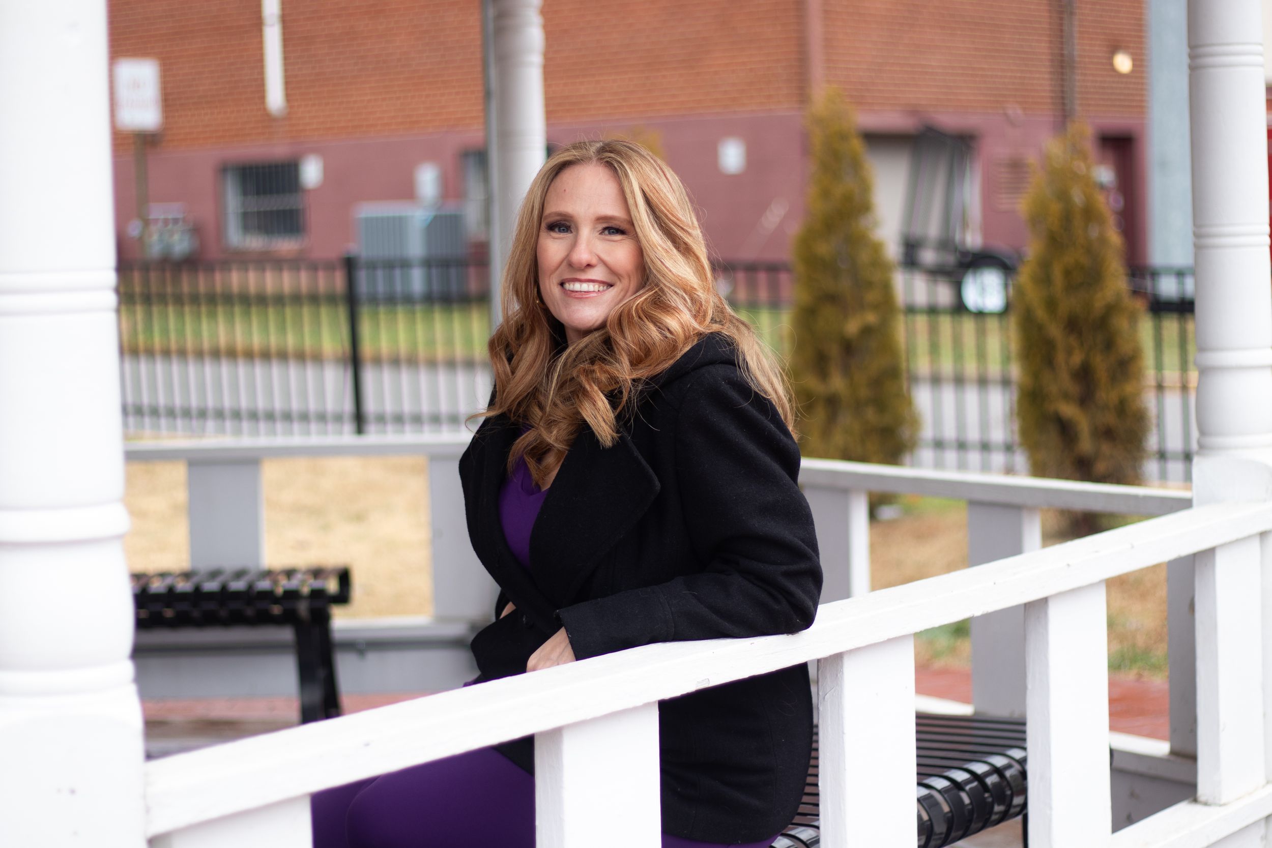 Person in black coat and purple dress posing by white railing outdoors.