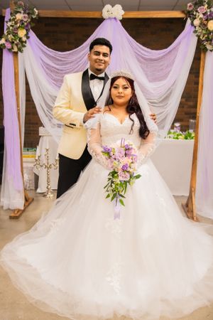 A couple poses under purple draping at their wedding with matching ivory formal wear.