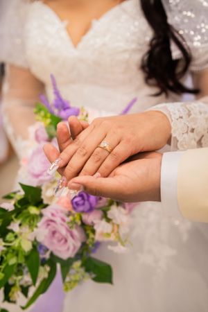 Close up of wedding rings and hands with purple floral bouquet in background.