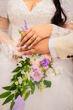 Close-up of hands with wedding rings resting on a bouquet of pink and purple roses with greenery.