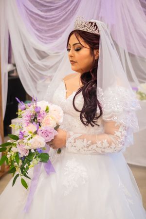 A beautiful bride in a white lace dress with long sleeves holds a purple and pink bouquet.
