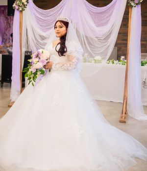 A stunning bride in a ballgown poses against a white and purple backdrop with floral accents.