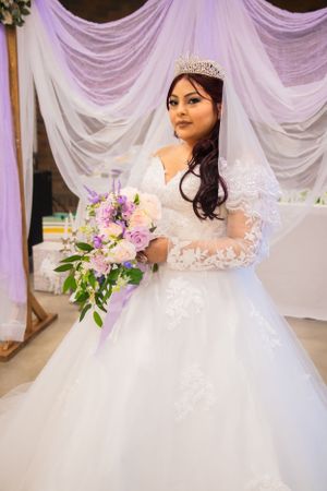 Elegant bride in white lace gown with crystal tiara holding purple floral bouquet.