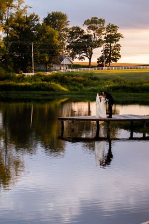 Scenic wedding photo with couple on dock during sunset with pastoral landscape backdrop.