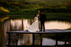 Couple in wedding attire striking a dramatic pose on lakeside dock at sunset.