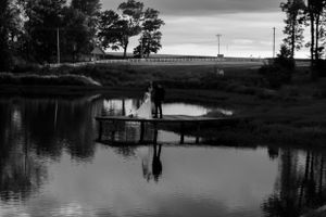 Black and white photo of silhouettes on a rustic dock over calm water at sunset.