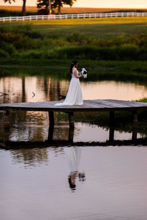 A person in a wedding dress stands on a wooden dock during golden hour with a white fence in background.
