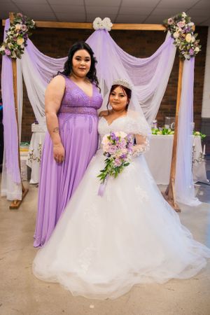 A bride in white poses with bridesmaid in purple under floral arch at wedding ceremony.