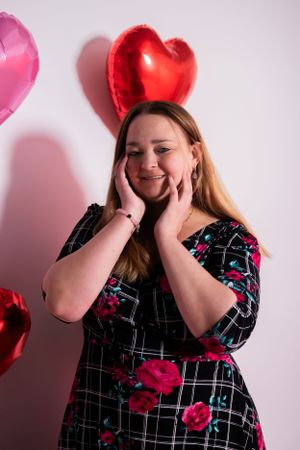 Person in floral dress posing with heart balloons against pink background.