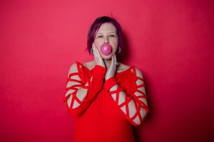 Person wearing red sweater with cutout sleeves blowing bubble gum against vibrant red background.