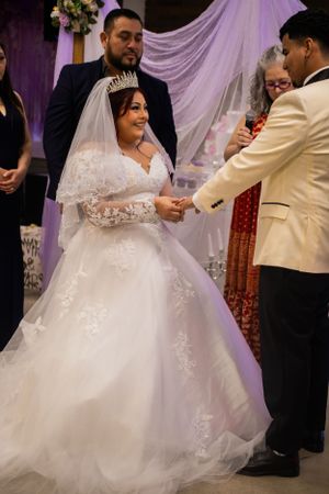 A bride in a lace gown exchanges rings during a wedding ceremony under purple draping and floral decor.