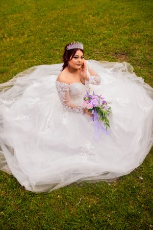 Aerial view of bride in white ballgown sitting on grass with purple bouquet.