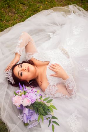 Bride in lace gown lying on grass with purple bouquet beside her.