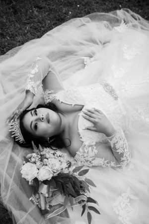 A bride in a lace wedding dress and tiara lies on tulle fabric with a bouquet of roses beside her.