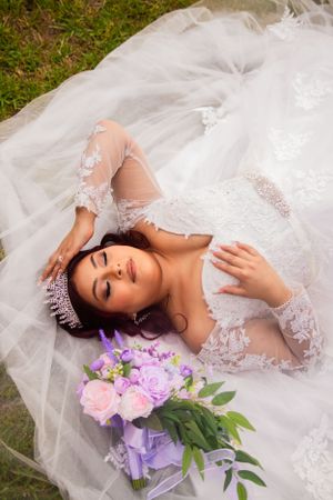 A bride in an embellished gown lies on tulle with a purple and pink floral bouquet.