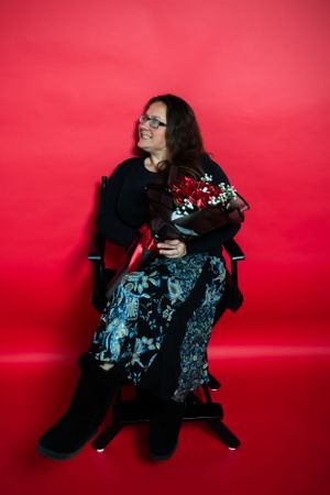 Person seated in black chair holding red rose bouquet against red backdrop.