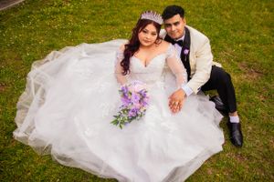 A newlywed couple sits on grass while wearing elegant white wedding attire with a purple bouquet.